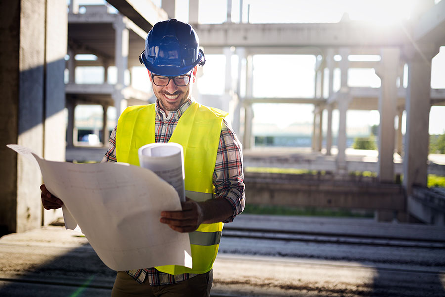 Specialized Business Insurance - Portrait of a Smiling Young Male Contractor Wearing a Hardhat Standing on a New Building Construction Jobsite While Holding Blueprints